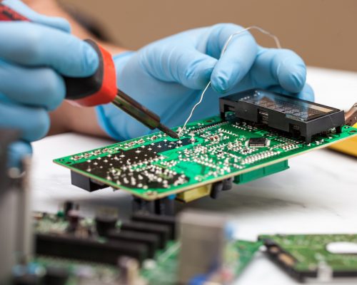 Hands wearing gloves solders and verifies computer circuit board using soldering iron and multimeter